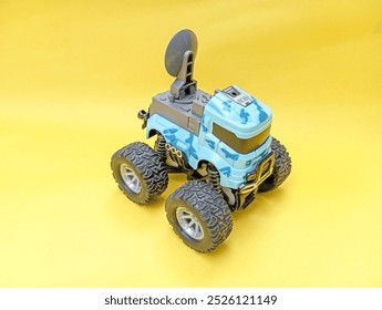 features a blue and gray toy monster truck with large, rugged wheels and a satellite dish mounted on top. The truck has a camouflage pattern and is positioned against a bright yellow background.