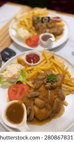 features a close-up, high-angle shot of two plates of chicken steak meals served on a white table. In the foreground, a white round plate showcases a tender, grilled chicken steak smothered in a light savory gravy and topped with sliced mushrooms and a fresh parsley leaf. The dish is accompanied by a generous serving of golden french fries, a small cup of tomato ketchup, a fresh slice of tomato, and a side of coleslaw salad resting on a green lettuce leaf. A small white gravy boat filled with additional brown sauce sits at the bottom left. In the blurred background, an identical second plate of the chicken steak meal is visible, creating a shallow depth-of-field effect that focuses on the front dish.