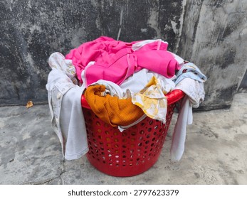 features a round red plastic laundry basket filled to the brim with a pile of dirty clothes or laundry ready to be washed. The pile is dominated by baby or children's clothing in various colors and patterns, including bright pink, white, mustard yellow, and light blue prints. The basket is placed outdoors against a backdrop of a weathered, textured gray concrete wall and a matching concrete floor.