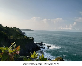 Features a scenic coastal landscape in Jogja, Indonesia. A rugged cliff covered in lush greenery dominates the left side of the frame, with terraced vegetation leading down towards the rocky shore. - Powered by Shutterstock