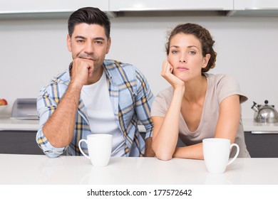 Fed Up Couple Having Coffee Looking At Camera At Home In Kitchen