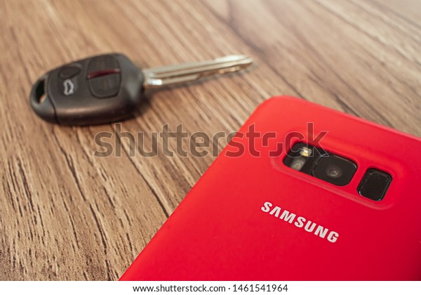 \
Brasília, Federal District - Brazil.\
July, 24, 2019. Mobile phone in a red colored cover with the name\
of samsung company and car key on a wooden\
table.