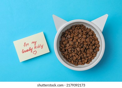 Feeding Bowl Of Kibble With Cat Ears And Cute Note For My Lovely Kitty On Light Blue Background, Flat Lay