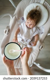 Feeding Small Caucasian Baby With Rice Cereal Mash Concept Hands Of Unknown Woman Mother Holding Bowl With Food Above Swing Or Feeding Seat With Baby Selective Focus On Hands And Spoon First Meal