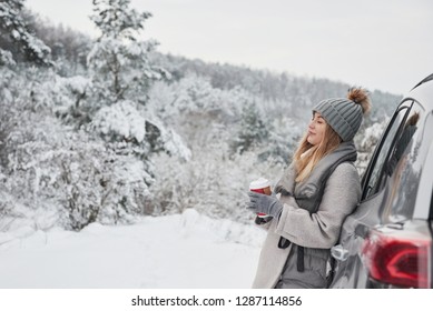 Feel The Moment. Pretty Girl In Warm Clothes Standing In The Winter Wood While Leans On The Car And Holding Cup Of Coffee.