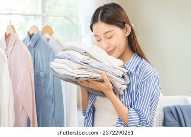 Feel softness, hygiene. Smile asian young woman touch fluffy white shirt smelling fresh clean pile, stack clothes, comfort sniff after washing laundry. Household work at home, chore of maid concept.