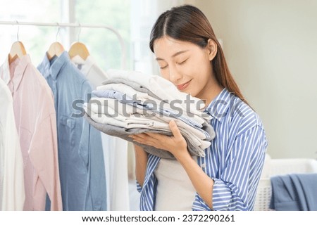 Feel softness, hygiene. Smile asian young woman touch fluffy white shirt smelling fresh clean pile, stack clothes, comfort sniff after washing laundry. Household work at home, chore of maid concept.