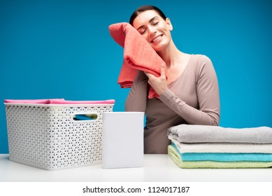 Feel softness. Smiling woman touching fluffy cotton towel with left cheek. She is enjoying nice smell with closed eyes. Wicker basket and washing powder are nearby