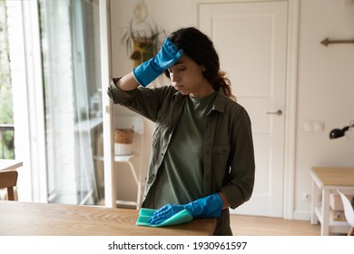 I feel for you poor Cinderella. Exhausted millennial housewife in blue latex gloves wipe sweat from brow. Sad fatigued young woman too tired of household chores cleaning flat house dusting furniture
