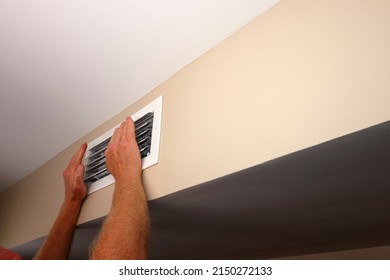 Feeling the air from a home air conditioning and heating HVAC vent on a wall are two hands in front of air duct grid. In a home near a ceiling is a rectangle white air vent with two hands in front of