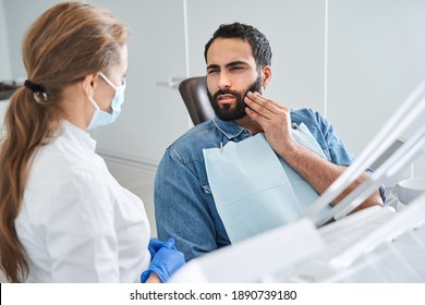Feeling bad. Young multiracial man having toothache, sitting in a dental chair at the clinic. Bearded male patient with terrible toothache visiting dentist. Stock photo