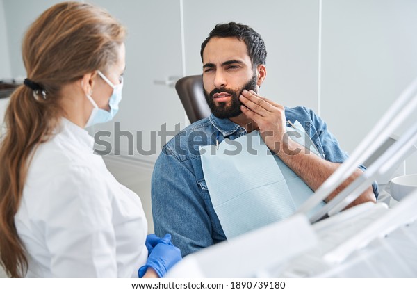 Feeling bad. Young\
multiracial man having toothache, sitting in a dental chair at the\
clinic. Bearded male patient with terrible toothache visiting\
dentist. Stock photo