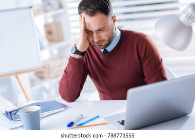 Feeling dizzy. Tired young unhappy man sitting at the table in his big office and touching his head while having strange feelings in it