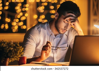 Feeling exhausted. Frustrated young man carrying eyeglasses and keeping eyes closed while sitting at his working place at night time with Christmas lights in the background - Powered by Shutterstock