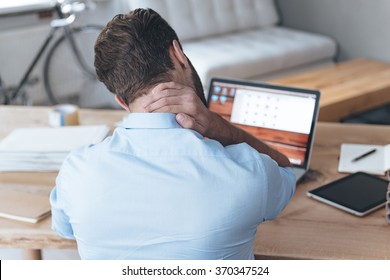Feeling exhausted. Rear view of frustrated young man looking exhausted and massaging his neck while sitting at his working place 