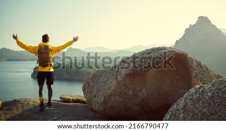 Feeling of freedom, achievement. A man tourist with a backpack behind his back stands on top of a mountain with outstretched arms, enjoying the scenery around. Active lifestyle, hiking.