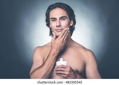Feeling fresh after shaving. Portrait of handsome young shirtless man looking at camera and applying aftershave lotion on face while standing against grey background 