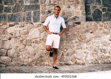 Feeling good and looking great. Full length of handsome young African man in smart casual clothes holding hands in pockets and smiling while leaning at the stoned wall outdoors 