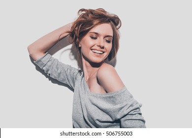 Feeling So Happy. Attractive Young Smiling Woman Holding Hand In Hair And Keeping Eyes Closed While Standing Against Grey Background