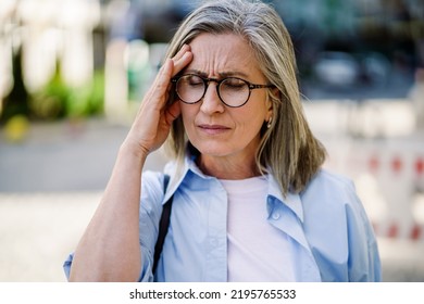 Feeling Hard Headache, Migraine Mature Grey Haired Woman 50s, 60s, Standing Outdoors Holding Her Head. Suffering From Summer Teat Feeling Sick While Travel Mature Woman Standing Outdoor. 