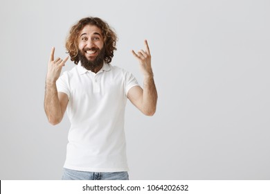 Feeling Like Rockstar. Portrait Of Upbeat Positive Eastern Guy With Beard And Curly Hair Showing Rock Gestures And Smiling Broadly, Feeling Great Attending Concert Or Making Deal And Celebrating It