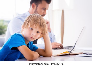 Feeling so lonely. Sad little boy holding hand in hair and looking at camera while his busy father working in the background 