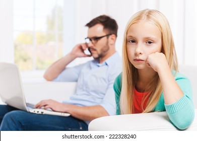 Feeling Lonely At Those Moments. Busy Young Man Working On Laptop And Talking On The Mobile Phone With His Daughter Sitting On Foreground And Looking Upset 