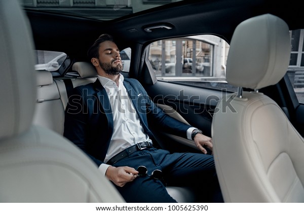 Feeling overworked. Tired young man in\
full suit keeping eyes closed while sitting in the\
car