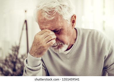 Feeling Pain. Old Sick Grey-haired Man In Beige Sweater Feeling Terrible Pain And Keeping His Hand On The Nose Bridge