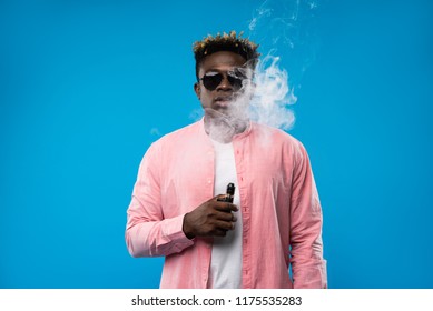 Feeling relaxed. Waist up portrait of afro guy holding vape device and surrounded by cloud of smoke. He is wearing pink shirt and sunglasses. Isolated on blue background