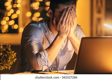 Feeling sick and tired. Frustrated young man covering face with hands while sitting at his working place at night time with Christmas lights in the background - Powered by Shutterstock