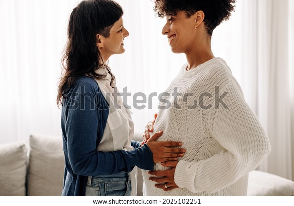 Feeling a\
surrogate mother\'s belly bump. Happy young woman smiling while\
feeling the movement of a pregnant woman\'s baby. Young woman\
spending time with her surrogate at\
home.