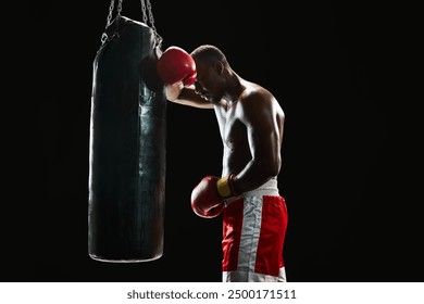 Feeling tired after intensive training. Muscular African man with strong body, boxer leaning on punching bag isolated on black background. Concept of professional sport, active lifestyle, strength - Powered by Shutterstock