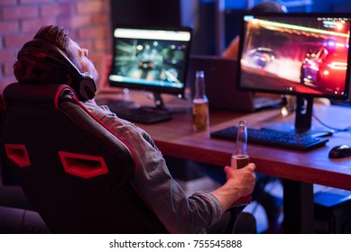 Feeling Tired. Back View Of Relaxed Young Gamer In Headphones Is Resting In Chair And Drinking Beer While Enjoying Car Racing Video Game. Selective Focus