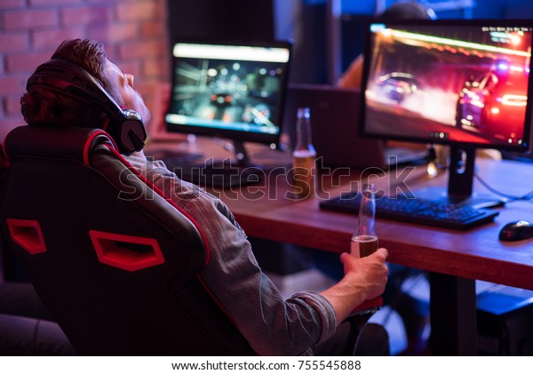 Feeling tired. Back view of relaxed young\
gamer in headphones is resting in chair and drinking beer while\
enjoying car racing video game. Selective\
focus