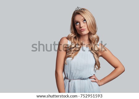 Feeling uncertain. Prideful young woman in crown keeping hand on hip and looking away while standing against grey background