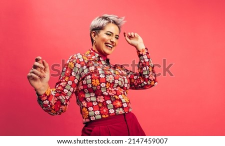 Feeling vibrant and energetic. Happy young woman dancing and smiling cheerfully while standing against a red background. Young woman with dyed hair having fun in the studio.