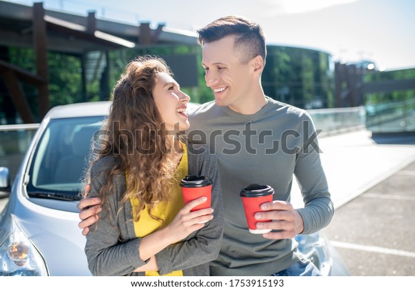 Feelings, love.\
Young adult man and woman with coffee standing near a car, looking\
at each other in love\
smiling.