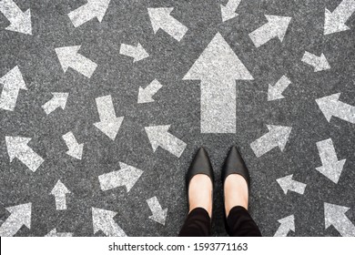Feet and arrows on road background from above. Businesswoman standing on pathway with drawn white many direction arrows choice. Top view of A businesswoman black shoes. Motivation and growth concept.