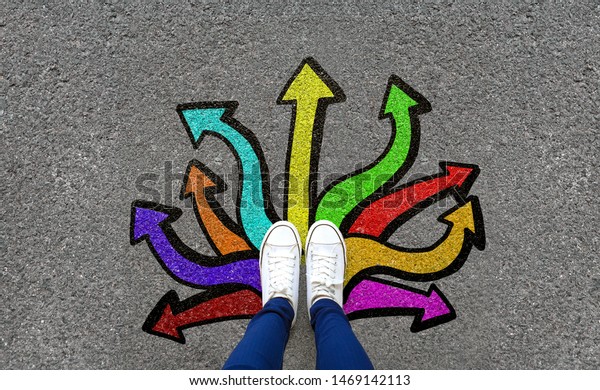 Feet and arrows on road background. Pair of foot\
standing on tarmac road with colorful graffiti arrow sign choices,\
creative and idea concept. Selfie woman wearing white shoe or\
sneaker. Top view.