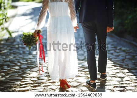 The feet of the bride and the groom with wedding bouquet. Bride in an elegant white dress and red shoes holds a stylish wedding bouquet with red and burgundy colors in her hands. Wedding details. 