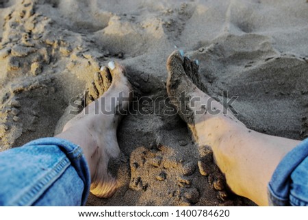  Feet buried in sand at the beach. Long legs in ripped blue jeans. Foot with blue nail varnish in wet sand. Young woman photographed from above sitting on the cold sand.