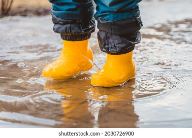 The Feet Of A Child In Yellow Rubber Boots Stomp Through Puddles