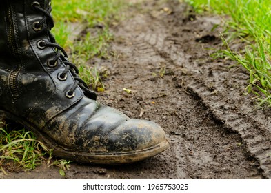 The Feet Of A Man In Boots On Muddy Country Road. Dirty Ground. Dirty Army Boots Walking On The Rainy Road. Spring. Autumn. Agriculture Details - Country Outdoor Scenery.