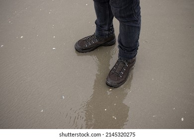 Feet Of A Man In Hiking Shoes Standing On A Seashore. Hiking Boots, Water Resistance Concept.