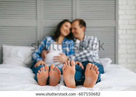 Feet of man and woman on bed with white linen in bedroom. Pregnant woman and man lying in the bedroom. Happy husband takes hands on wifes belly. Waiting for a baby. Family expecting for a baby.