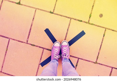 Feet With Purple Tennis Shoes Standing On A Black Cross On A Pavement, Colourful Background