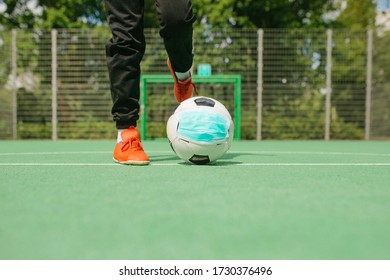 Feet And Shoes Of Boy Kicking Off Soccer Football With Surgical Face Mask On Artifiical Turf Field, Symbol Image For Sports During The Corona Covid-19 Pandemic In 2020