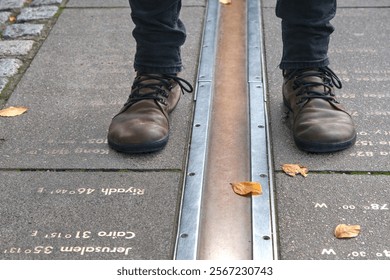 Feet straddling the Prime Meridian at Greenwich, London, with longitude markers engraved on the ground, representing global time zones.