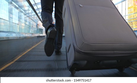 Feet Of Successful Businessman Walking In Hall Of Terminal With His Luggage. Legs Of Wealthy Businessman Walking Escalator And Pulling Suitcase On Wheels. Concept Of Business Trip. Slow Mo Back View